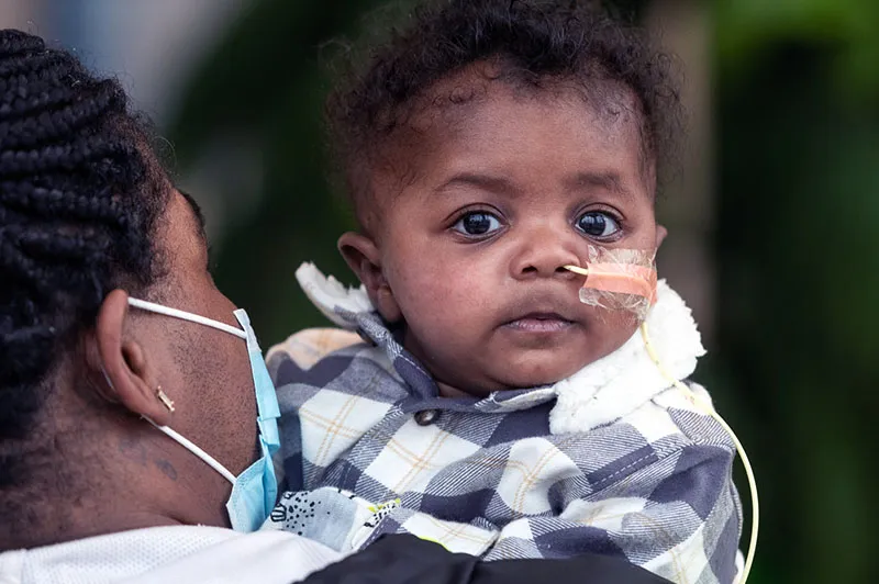 Wide-eyed child wearing a jacket is held by woman wearing a jacket and a face mask.