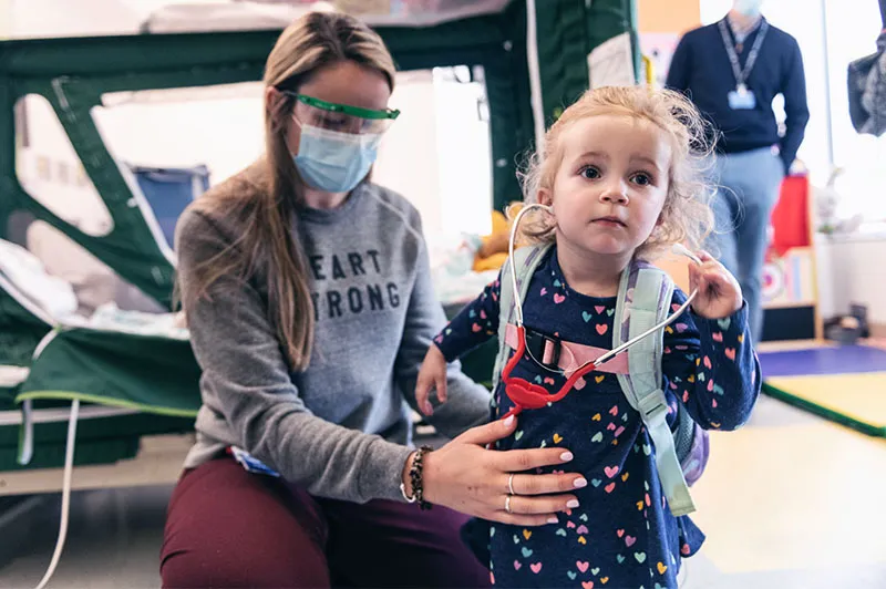 Young girl stands up, while being assisted by woman in sweatpants and face mask.