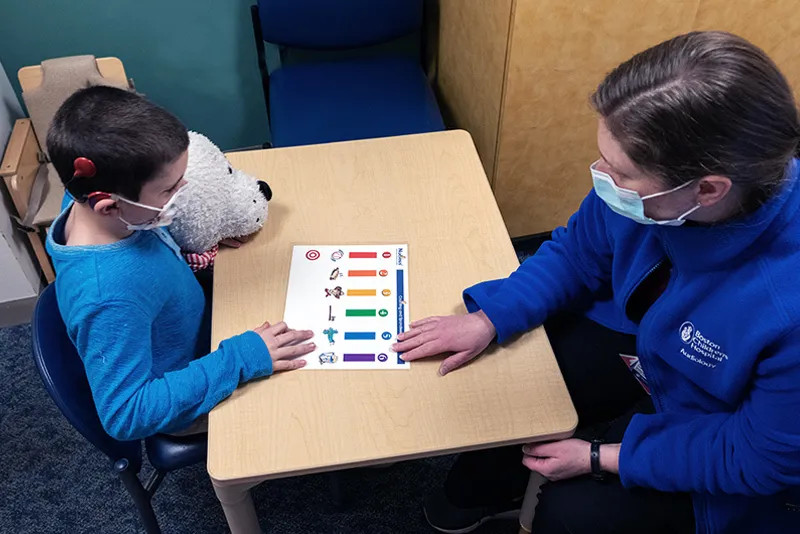 Clinician on right wearing blue jacket and face mask works with young boy on speech