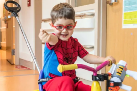Boy wearing Spider-Man outfit rides on small tricycle through hospital unit